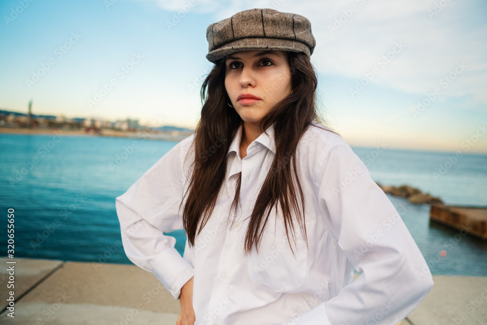 Portrait of young beautiful stylish woman in white shirt and grey cap. She is on terrace with ocean view