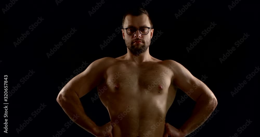 Close-up portrait of a dark-haired man with glasses, he is in the Studio on a black background. His torso is bare, and he straightens as much as possible, pulling his shoulders back. Close-up portrait of a dark-haired man with glasses, he is in the Studio on a black background. His torso is bare, and he straightens as much as possible, pulling his shoulders back.