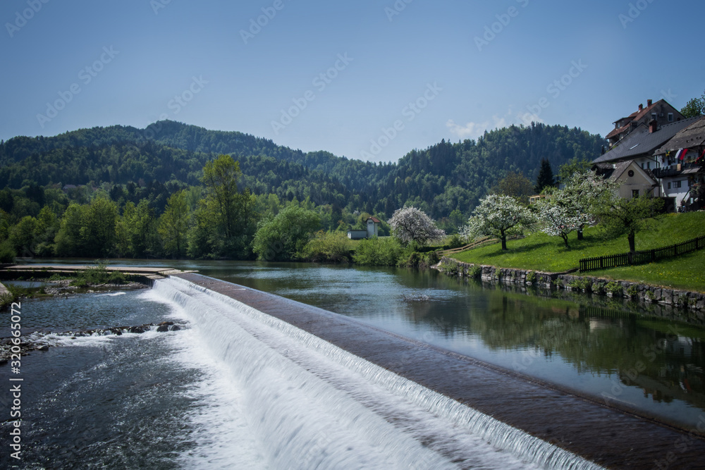 A small hand made water dam on a small river of Sora, Slovenia on a sunny day. Background with small green hills.