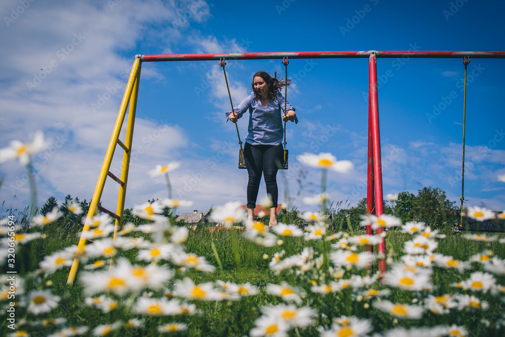 Young brunette woman is swinging on a swing, with a field of daisies (anthemidae) in the foreground on a beautiful spring sunny day. Concept of natural playfulness outdoor. Young brunette woman is swinging on a swing, with a field of daisies (anthemidae) in the foreground on a beautiful spring sunny day. Concept of natural playfulness outdoor.