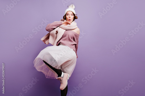 Studio shot of stunning caucasian woman wears winter hat and lush skirt. Portrait of adorable girl in fashionable attire isolated on purple background.