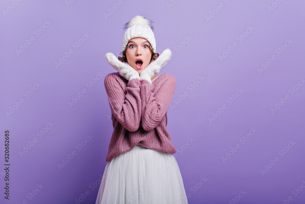 Amazed caucasian girl in winter outfit posing in studio with purple interior. Surprised young woman in white skirt and knitted sweater standing with mouth open.