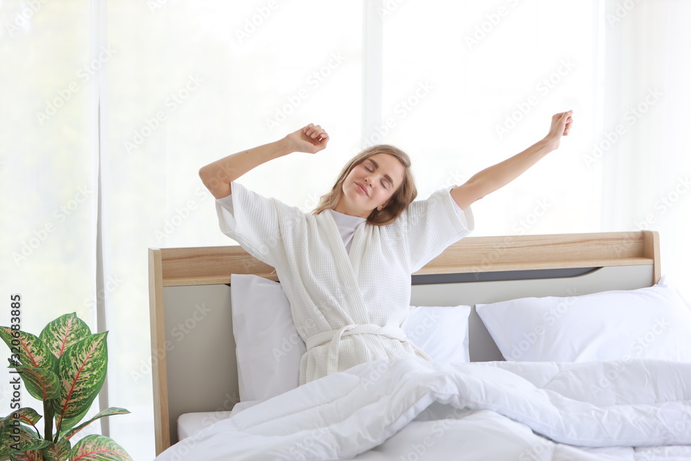 Cheerful and healthy caucasian young woman wearing pajama wake up in the morning at sunrise and sitting and stretching on bed in bedroom with relaxation and happy face Cheerful and healthy caucasian young woman wearing pajama wake up in the morning at sunrise and sitting and stretching on bed in bedroom with relaxation and happy face