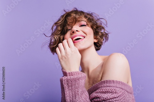 Cheerful white woman with playful smile posing in soft knitted sweater. Indoor portrait of good-looking young lady with short haircut chilling on purple background.