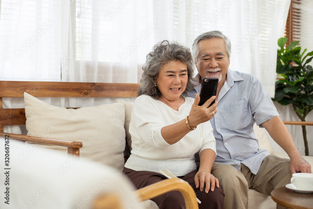 Happy Asian smile couple sitting on sofa using smartphone together video chatting with family in living room at home or selfie, concept of connected technology for communication, retirement lifestyle Happy Asian smile couple sitting on sofa using smartphone together video chatting with family in living room at home or selfie, concept of connected technology for communication, retirement lifestyle