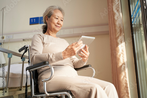 asian old woman sitting in wheel chair looking at digital tablet in nursing home or hospital ward photo