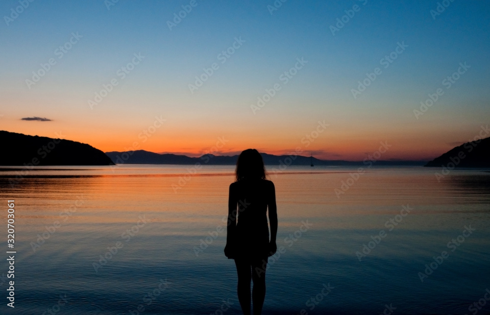 black silhouette of a woman on the background of a stunning orange and blue sunset on the sea, the sunset sky is reflected in the water