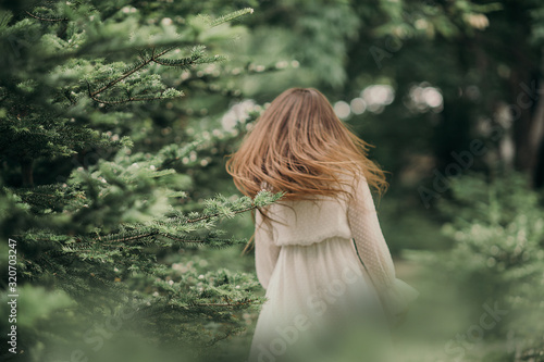 green prickly branches of a tree in focus against the background of a fleeing blurry girl with long straight hair and a white cotton dress out of focus