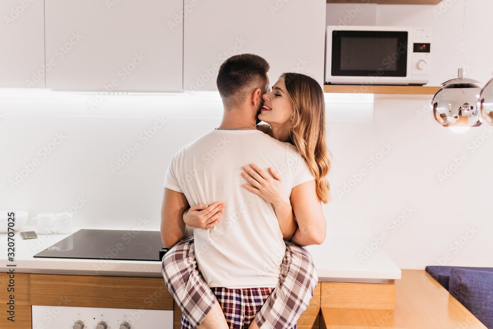 Blithesome girl in stylish pajama embracing husband with sincere smile. Caucasian married couple posing in kitchen. Blithesome girl in stylish pajama embracing husband with sincere smile. Caucasian married couple posing in kitchen.