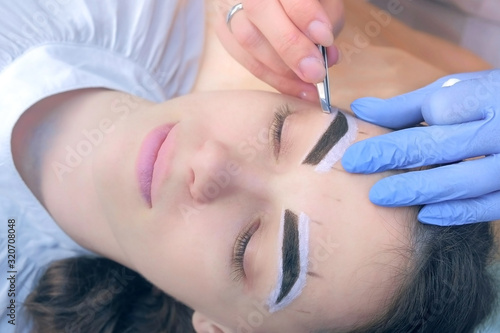 Professional beautician plucking eyebrows with tweezers to woman in beauty salon during tint eyebrow procedure  face closeup. Girl lying with closed eyes and brown natural henna on brows.