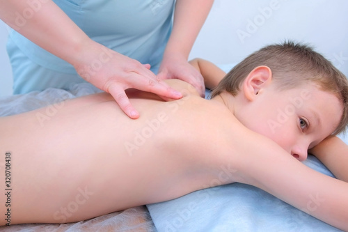Doctor massagist making therapeutic massage to teen boy on spine in clinic, side view. Child boy is lying on couch and talking. Rehabilitation massage after injury. Medicine and treatment to children.