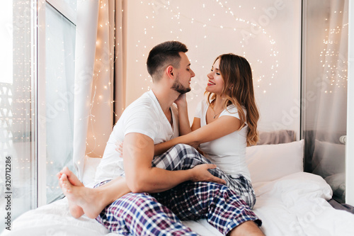 Barefooted lady in cozy pajama sitting on boyfriend's knees. Glad female model looking into husband's eyes.