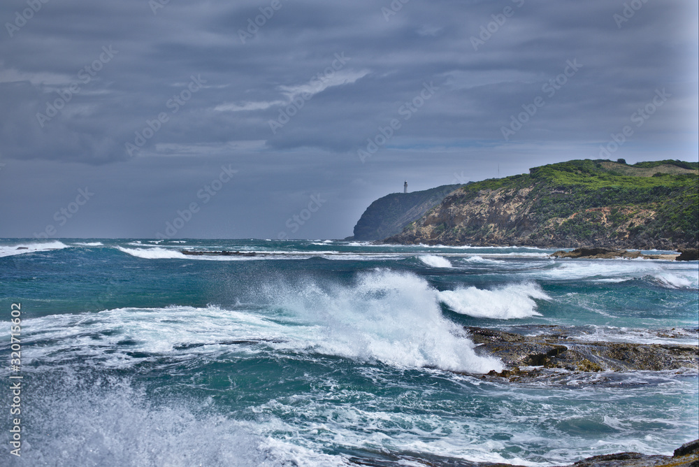 Landscape photo of rough seas with waves crashing in the foreground and green clifftops in the background one with a light house on it.