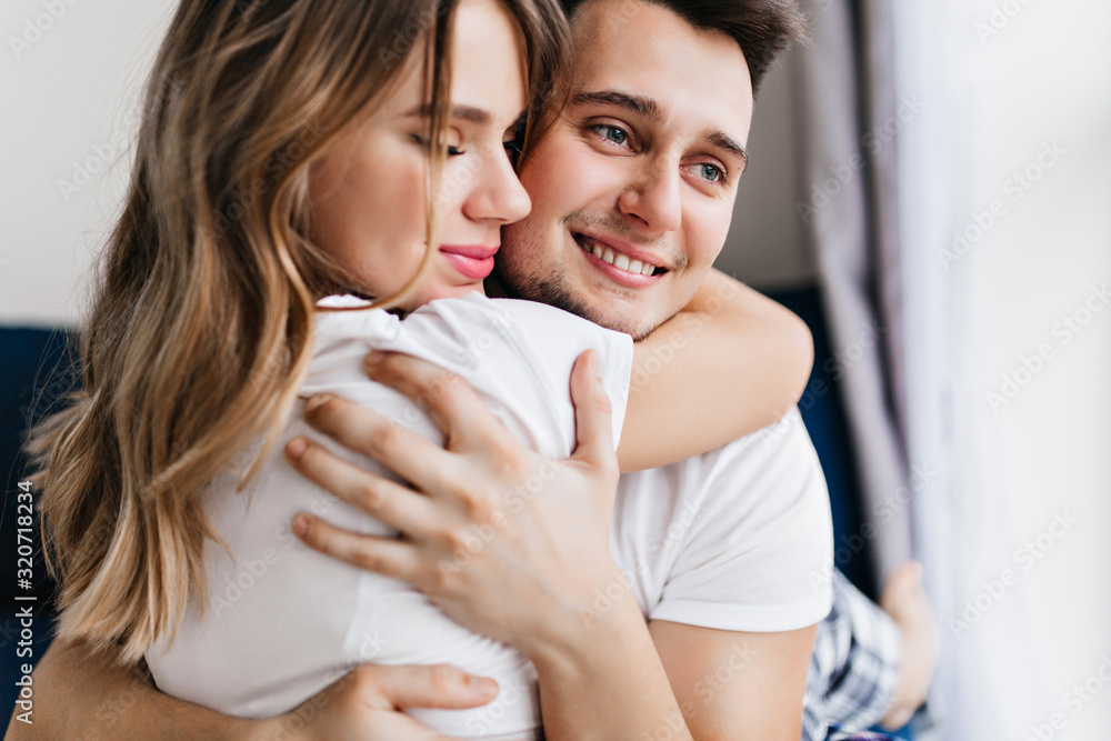 Blissful handsome man huggs his girlfriend. Indoor portrait of european married couple enjoying weekend morning. Blissful handsome man huggs his girlfriend. Indoor portrait of european married couple enjoying weekend morning.