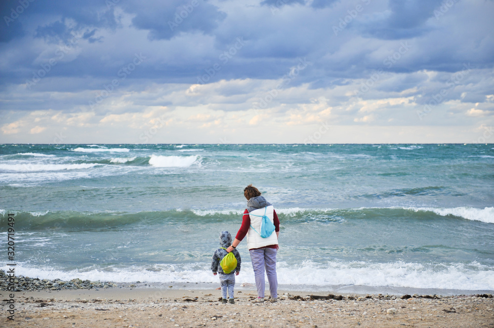adult woman grandmother together with little kid in hood and jacket with backpack during travel near stormy cold spring sea with moody sky adult woman grandmother together with little kid in hood and jacket with backpack during travel near stormy cold spring sea with moody sky