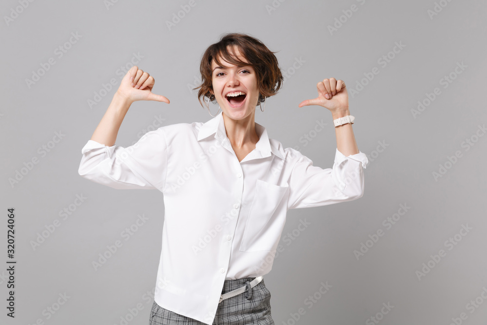 Cheerful funny young business woman in white shirt posing isolated on grey wall background studio portrait. Achievement career wealth business concept. Mock up copy space. Pointing thumbs on herself. Cheerful funny young business woman in white shirt posing isolated on grey wall background studio portrait. Achievement career wealth business concept. Mock up copy space. Pointing thumbs on herself.