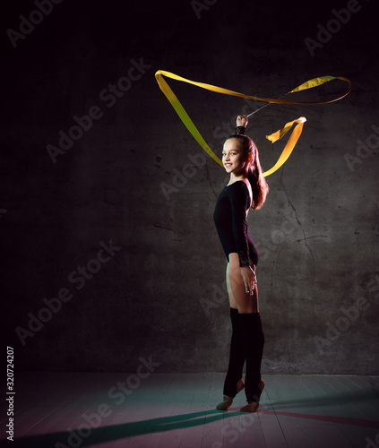 Young girl gymnast in black sport body and uppers standing with streched leg and making exercise with gymnastic tape © FAB.1