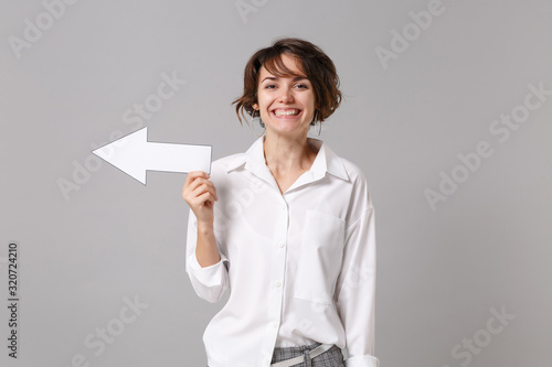 Smiling funny young business woman in white shirt posing isolated on grey wall background studio portrait. Achievement career wealth business concept. Mock up copy space. Showing aside with arrow,