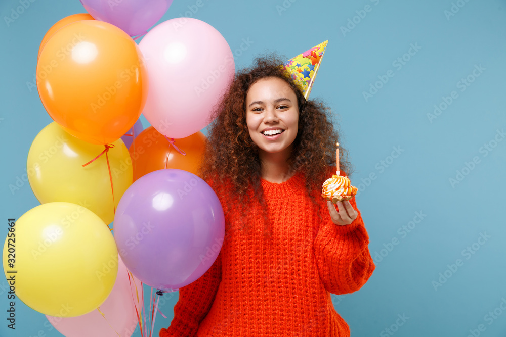 Obraz premium Smiling young african american girl in casual orange knitted clothes, birthday hat isolated on pastel blue background. Holiday party concept. Celebrating, hold colorful air balloons, cake with candle.