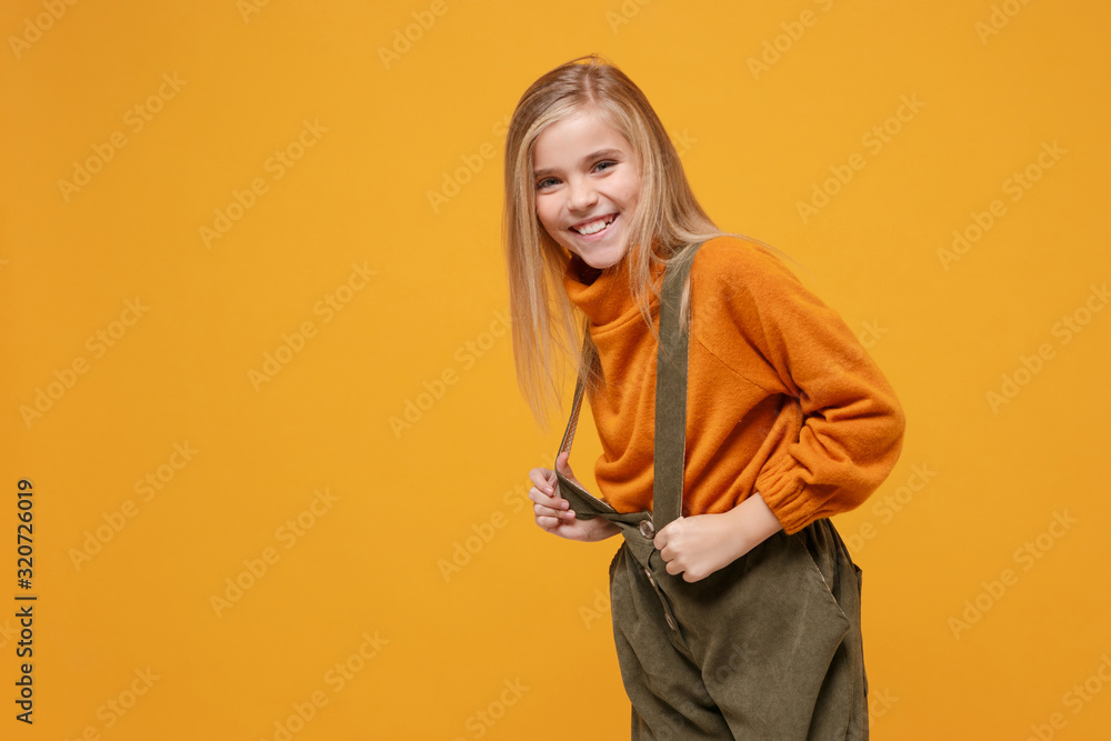 Charming little blonde kid girl 12-13 years old in turtleneck, jumpsuit isolated on orange yellow background children studio portrait. Childhood lifestyle concept. Mock up copy space. Looking camera. Charming little blonde kid girl 12-13 years old in turtleneck, jumpsuit isolated on orange yellow background children studio portrait. Childhood lifestyle concept. Mock up copy space. Looking camera.