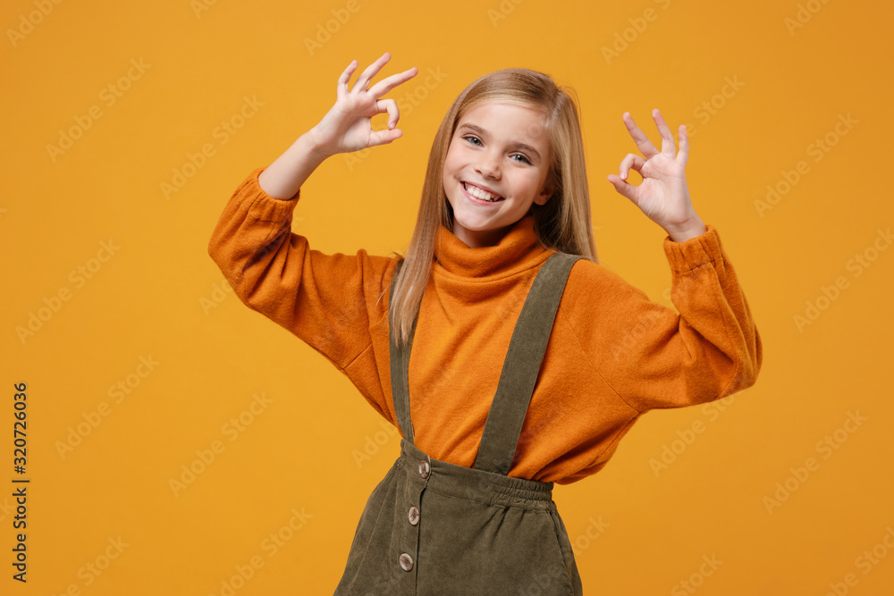 Smiling little kid girl 12-13 years old in turtleneck, jumpsuit isolated on orange yellow wall background children studio portrait. Childhood lifestyle concept. Mock up copy space. Showing OK gesture. Smiling little kid girl 12-13 years old in turtleneck, jumpsuit isolated on orange yellow wall background children studio portrait. Childhood lifestyle concept. Mock up copy space. Showing OK gesture.
