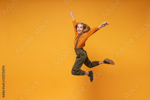 Funny little kid girl 12-13 years old in turtleneck, jumpsuit isolated on orange yellow wall background children studio portrait. Childhood lifestyle concept. Mock up copy space. Jumping rising hands.