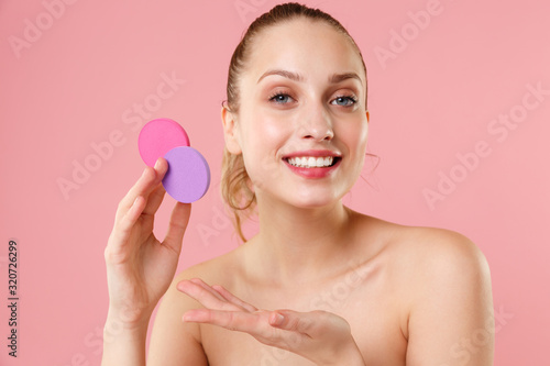 Close up blonde half naked woman 20s perfect skin blue eyes isolated on pastel pink wall background studio portrait. Skin care healthcare procedures concept Apply foundation nude makeup sponge on face