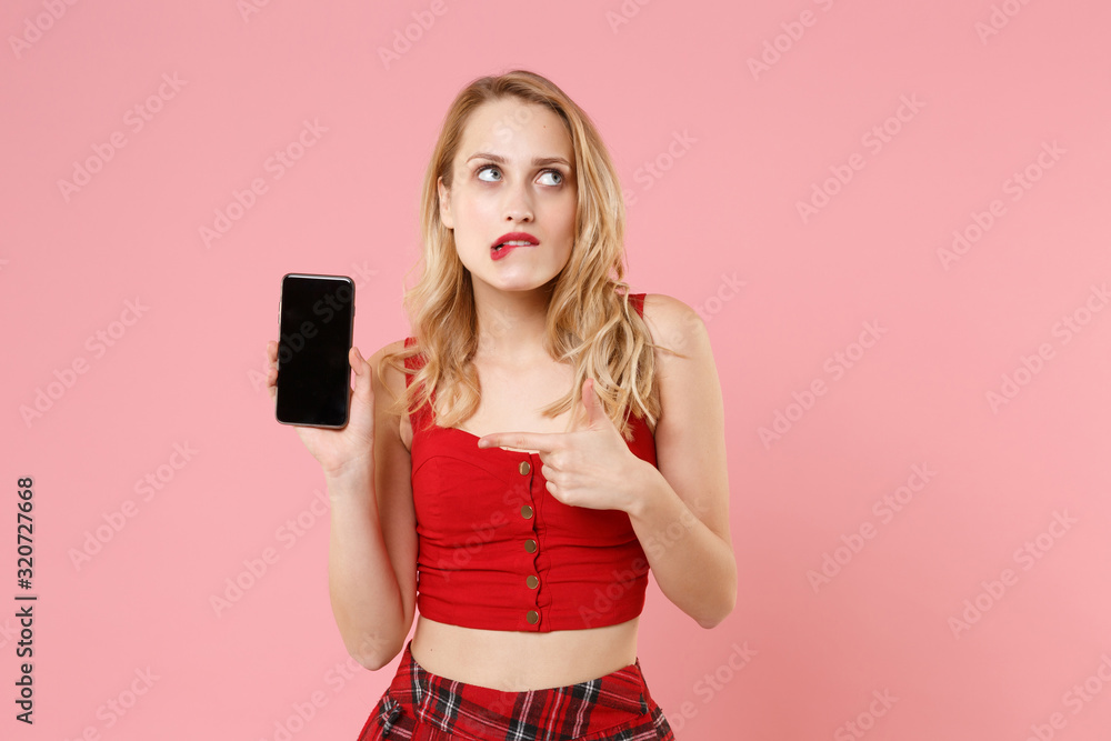 Confused young woman girl in red sexy clothes isolated on pastel pink background. People lifestyle concept. Mock up copy space. Point index finger on mobile phone with blank empty screen, biting lips. Confused young woman girl in red sexy clothes isolated on pastel pink background. People lifestyle concept. Mock up copy space. Point index finger on mobile phone with blank empty screen, biting lips.
