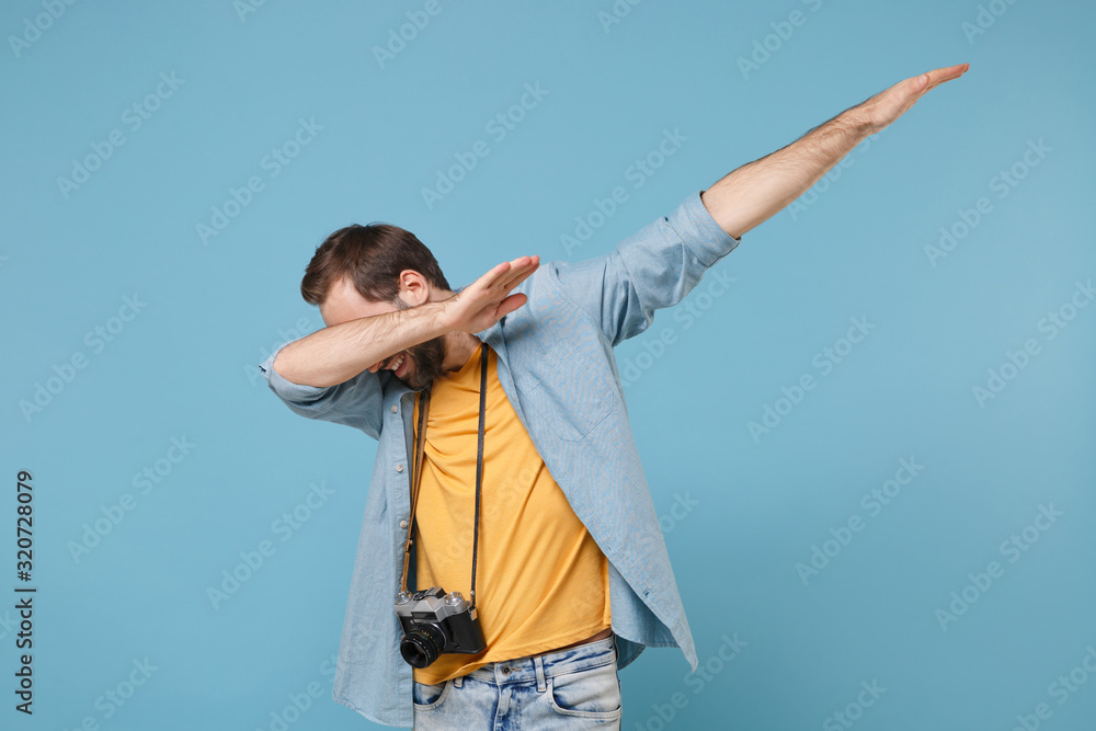Traveler tourist man in yellow summer casual clothes with photo camera isolated on blue background. Male passenger traveling abroad on weekends. Air flight journey concept. Showing DAB dance gesture. Traveler tourist man in yellow summer casual clothes with photo camera isolated on blue background. Male passenger traveling abroad on weekends. Air flight journey concept. Showing DAB dance gesture.