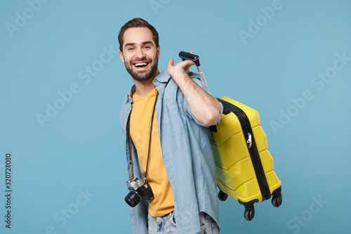 Cheerful traveler tourist man in yellow summer casual clothes with photo camera isolated on blue background. Male passenger traveling abroad on weekends. Air flight journey concept. Holding suitcase. photo
