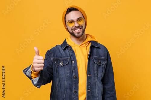 Smiling young hipster guy in fashion jeans denim clothes posing isolated on yellow orange background studio portrait. People sincere emotions lifestyle concept. Mock up copy space. Showing thumb up.