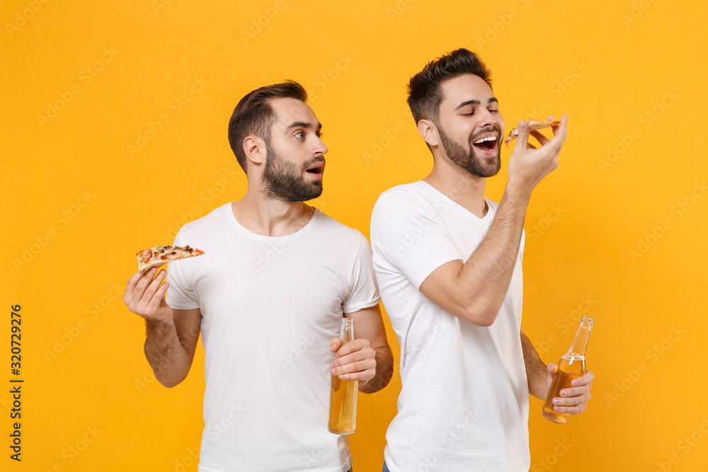 Amazed young men guys friends in white blank empty t-shirts posing isolated on yellow orange background. People sincere emotions lifestyle concept. Mock up copy space. Hold beer bottles italian pizza. Amazed young men guys friends in white blank empty t-shirts posing isolated on yellow orange background. People sincere emotions lifestyle concept. Mock up copy space. Hold beer bottles italian pizza.