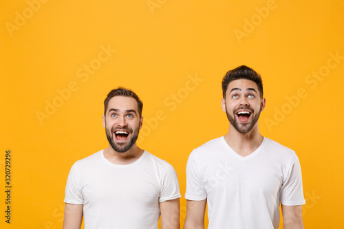 Excited young men guys friends in white blank empty t-shirts posing isolated on yellow orange background studio portrait. People lifestyle concept. Mock up copy space. Keeping mouth open, looking up.