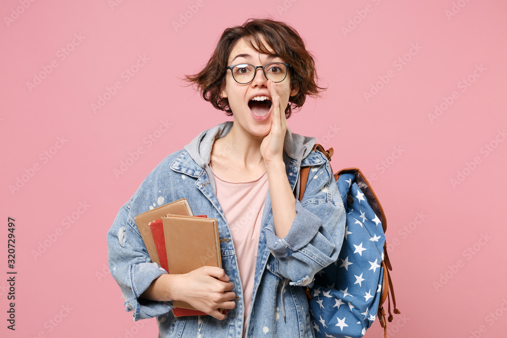 Young woman student in denim clothes eyeglasses backpack posing isolated on pastel pink background. Education in high school university college concept. Hold books whispering secret behind her hand. Young woman student in denim clothes eyeglasses backpack posing isolated on pastel pink background. Education in high school university college concept. Hold books whispering secret behind her hand.
