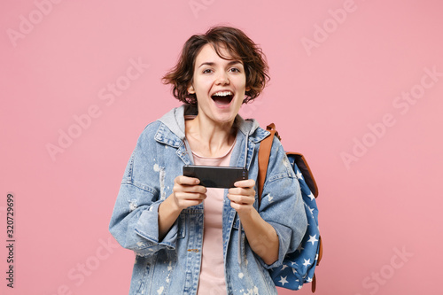 Cheerful young girl student in denim clothes backpack isolated on pastel pink background in studio. Education in high school university college concept. Mock up copy space Play game with mobile phone.
