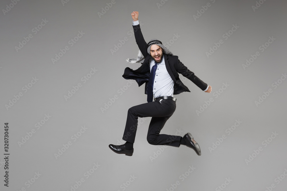 Cheerful bearded arabian muslim businessman in keffiyeh kafiya ring igal agal classic suit isolated on gray background. Achievement career wealth business concept. Jumping clenching fists like winner.