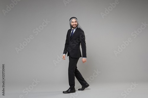 Smiling bearded young arabian muslim businessman in keffiyeh kafiya ring igal agal classic black suit shirt tie isolated on gray background. Achievement career wealth business concept. Looking aside.