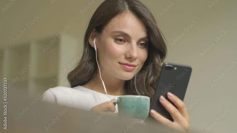 A nice calm woman with wired headphones is using her smartphone while drinking coffee indoors A nice calm woman with wired headphones is using her smartphone while drinking coffee indoors