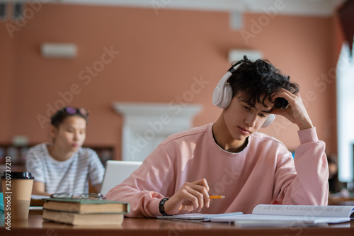 Bild auf Leinwand Pensive teenage student with headphones looking through page of manual before re