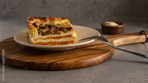 Baked vegetarian pumpkin lasagne on a plate and round wooden board. There is fork in a plate ant little bowl of cheese near the board. Grey background. 