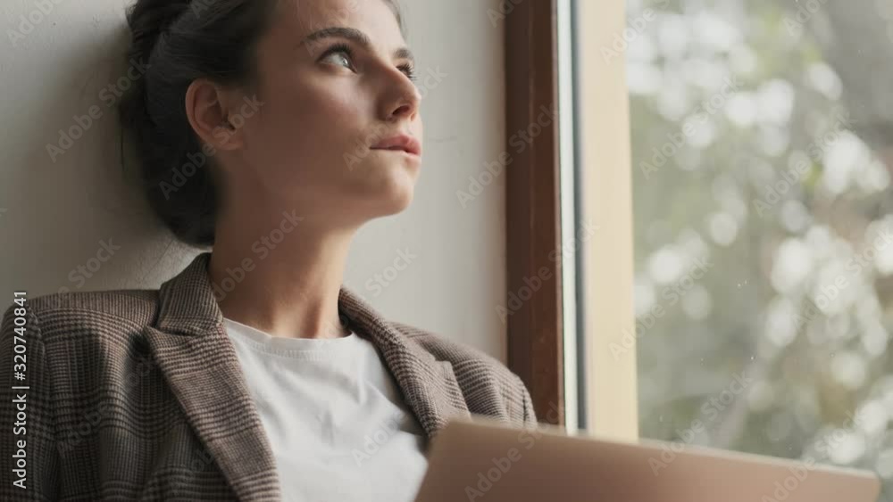 A thoughtful young woman is holding a laptop while sitting near the window A thoughtful young woman is holding a laptop while sitting near the window