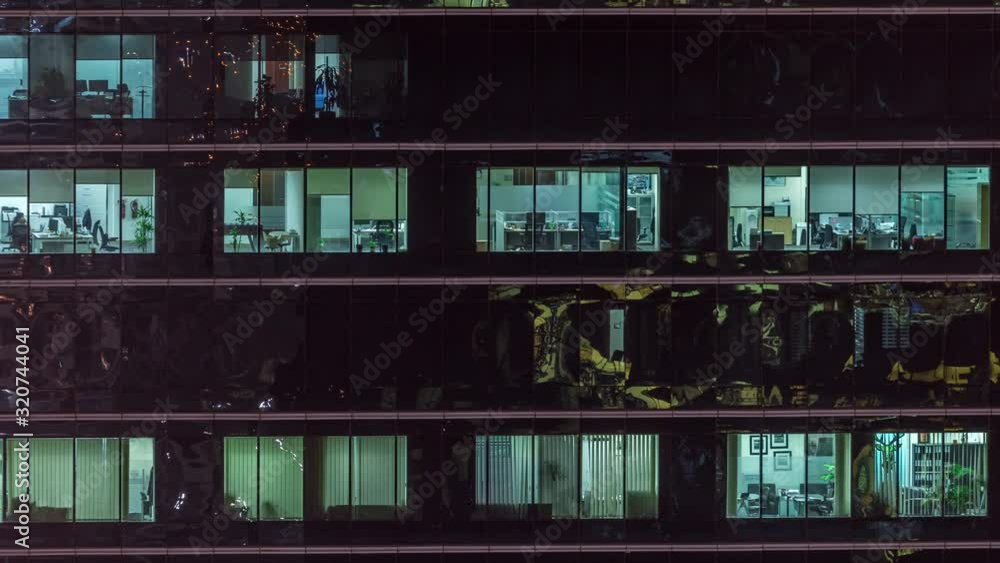 Office skyscraper exterior during late evening with interior lights on and people working inside night timelapse. Aerial close up view from above with many windows. Pan right Office skyscraper exterior during late evening with interior lights on and people working inside night timelapse. Aerial close up view from above with many windows. Pan right