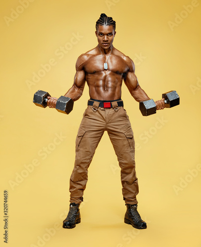 Man doing the exercises with dumbbells. Photo of young man with naked torso on yellow background. Strength and motivation