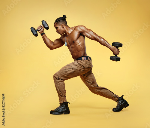 Sporty man training muscles of hands and legs using a dumbbells. Photo of man with good physique isolated on yellow background. Strength and motivation © Romario Ien