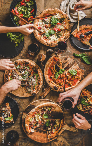 Friends having pizza party dinner. Flat-lay of people eating different kinds of Italian pizza, salad and drinking red wine over wooden table, top view. Fast food lunch, gathering, celebration