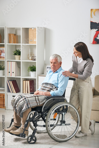 Young female care giver talking to grey-haired senior male pensioner sitting in wheelchair while staying at home