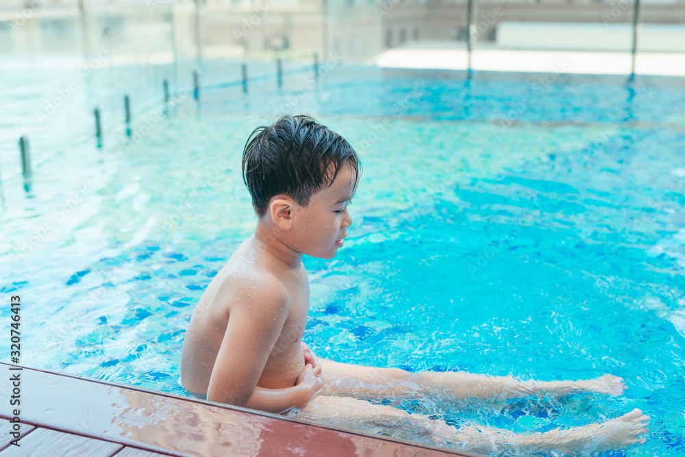 Asian cute child swimming in swimming pool. He playing is funny on summer holiday on blurred background. Cute boy smile and swim on daytime and light of sun. It joyful vacation for him.