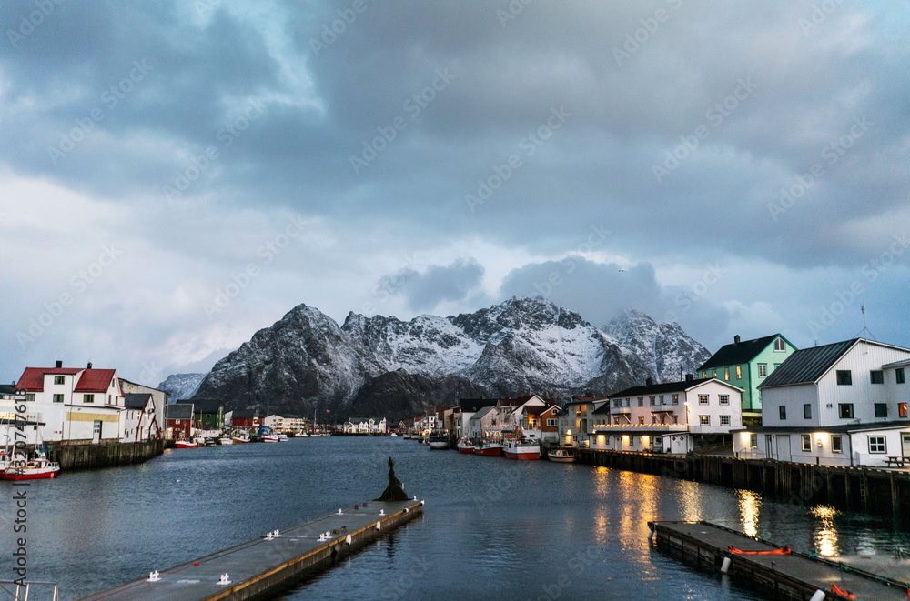 Henningsvaer harbor photo with buildings reflecting in water, mountains and overcast sky on horizon.