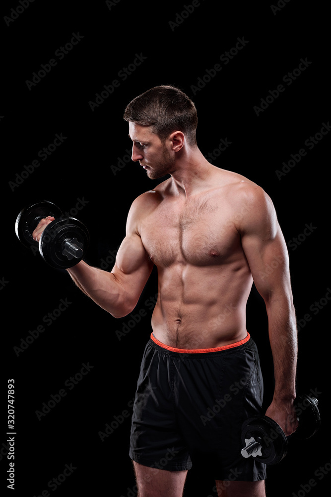 Young muscular shirtless weightlifter in shorts doing exercise for arms with dumbbells over black background