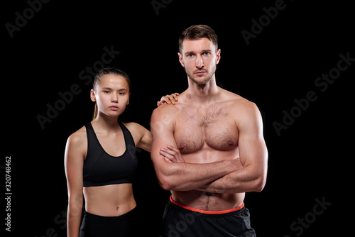 Handsome muscular shirtless guy and his fit pretty girlfriend in activewear standing close to one another on dark background